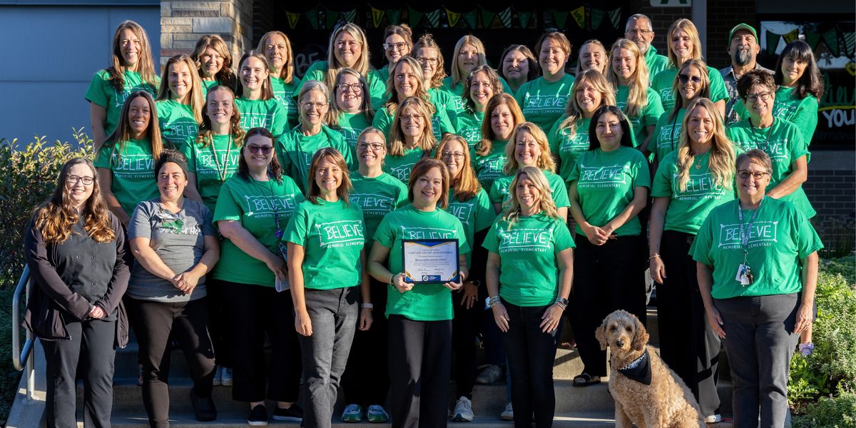 About three dozen Memorial Elementary staff members in matching green BELIEVE shirts pose on the school's front steps while a woman in the front row holds a framed award