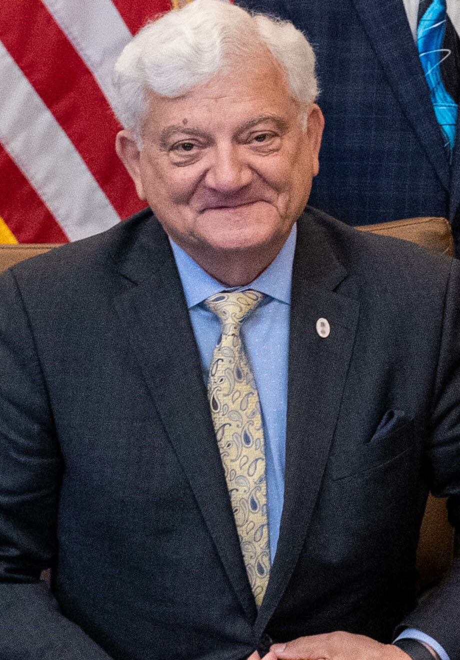 Portrait of state Rep. Ed Soliday in a dark suit and patterned tie, seated in front of an American flag.