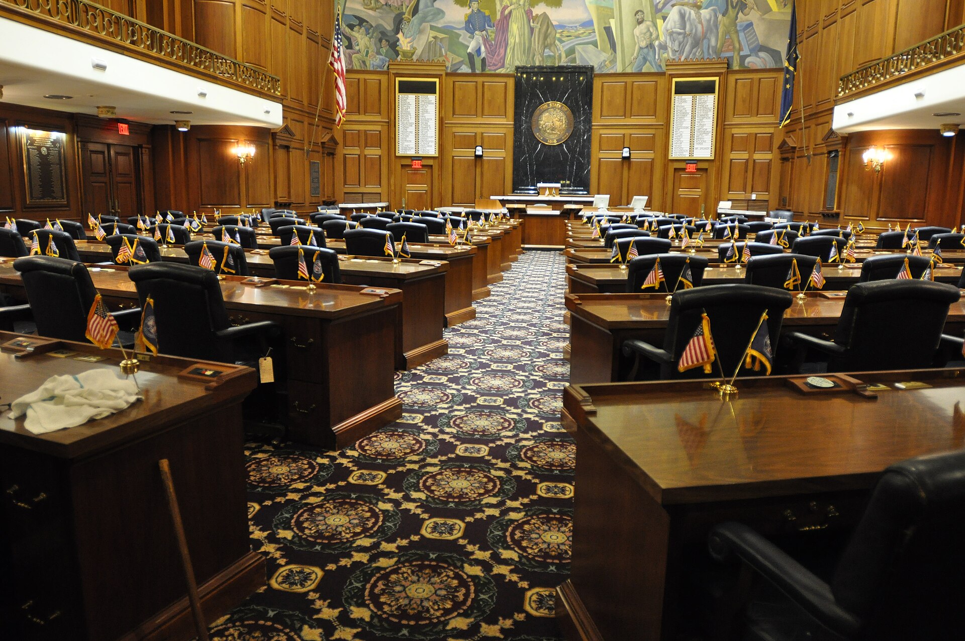The empty Indiana House of Representatives chamber at the Statehouse, with rows of wooden desks and small flags.