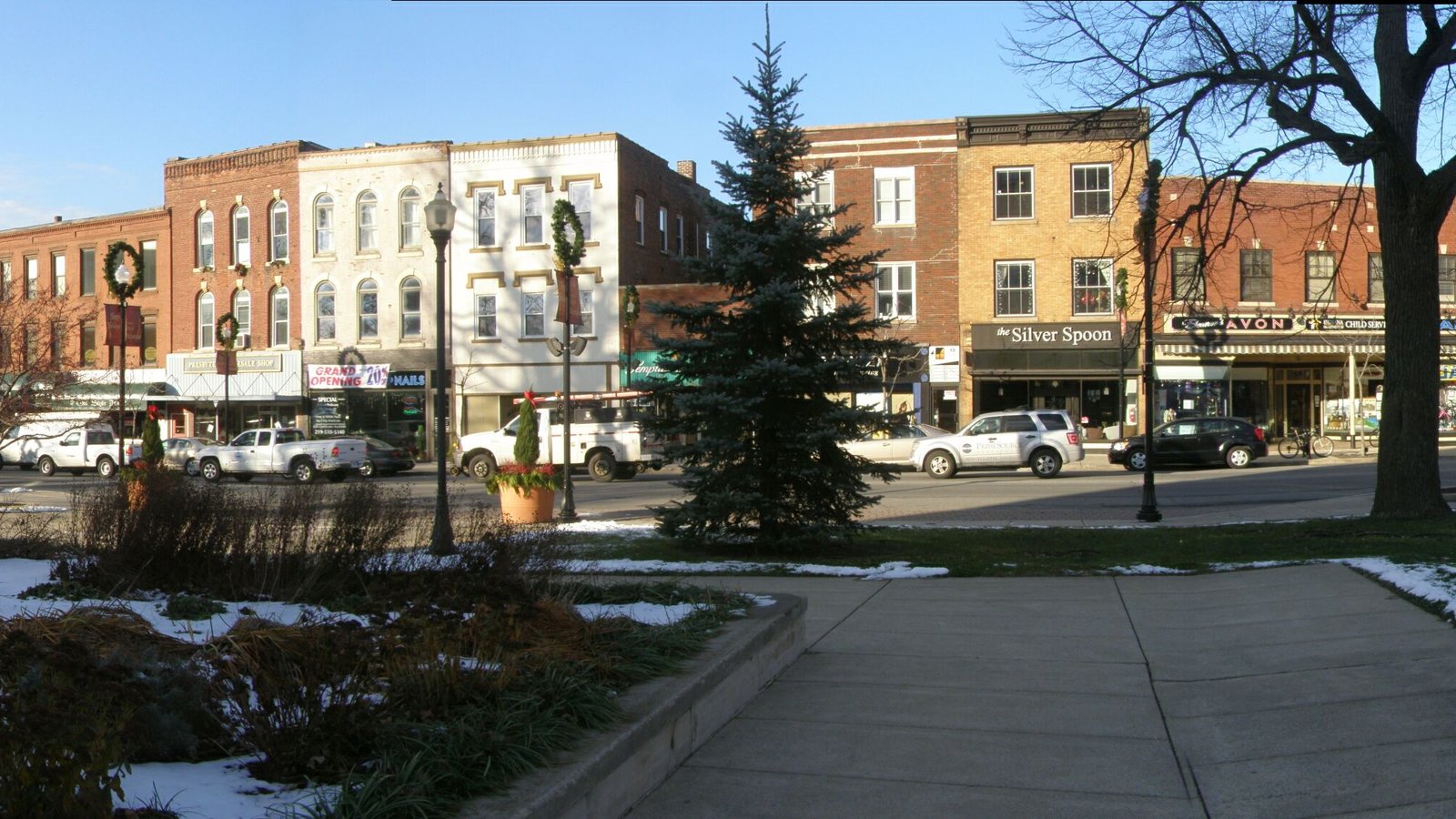 Row of two- and three-story brick storefronts across a street from a courthouse square lawn with an evergreen tree and patches of snow