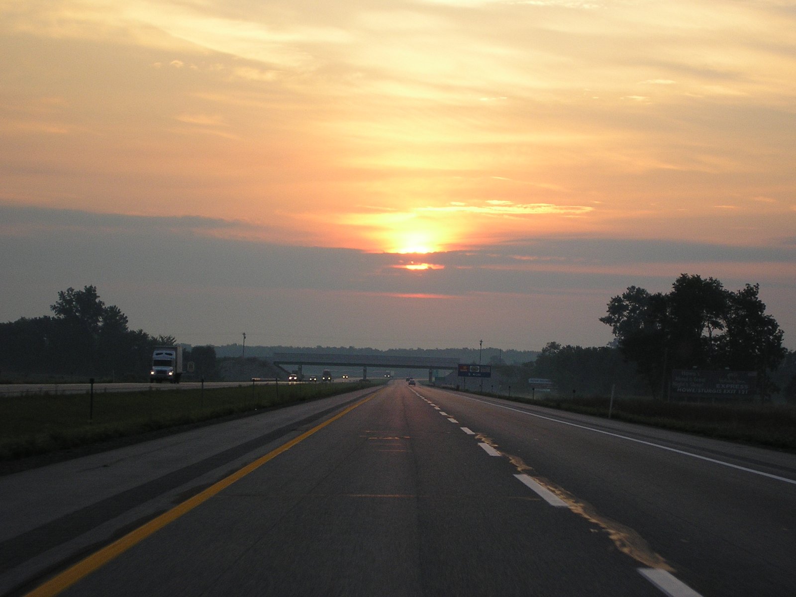 A divided four-lane interstate stretching toward an overpass under an orange sky with a low sun, with a semi-truck in the opposite lanes