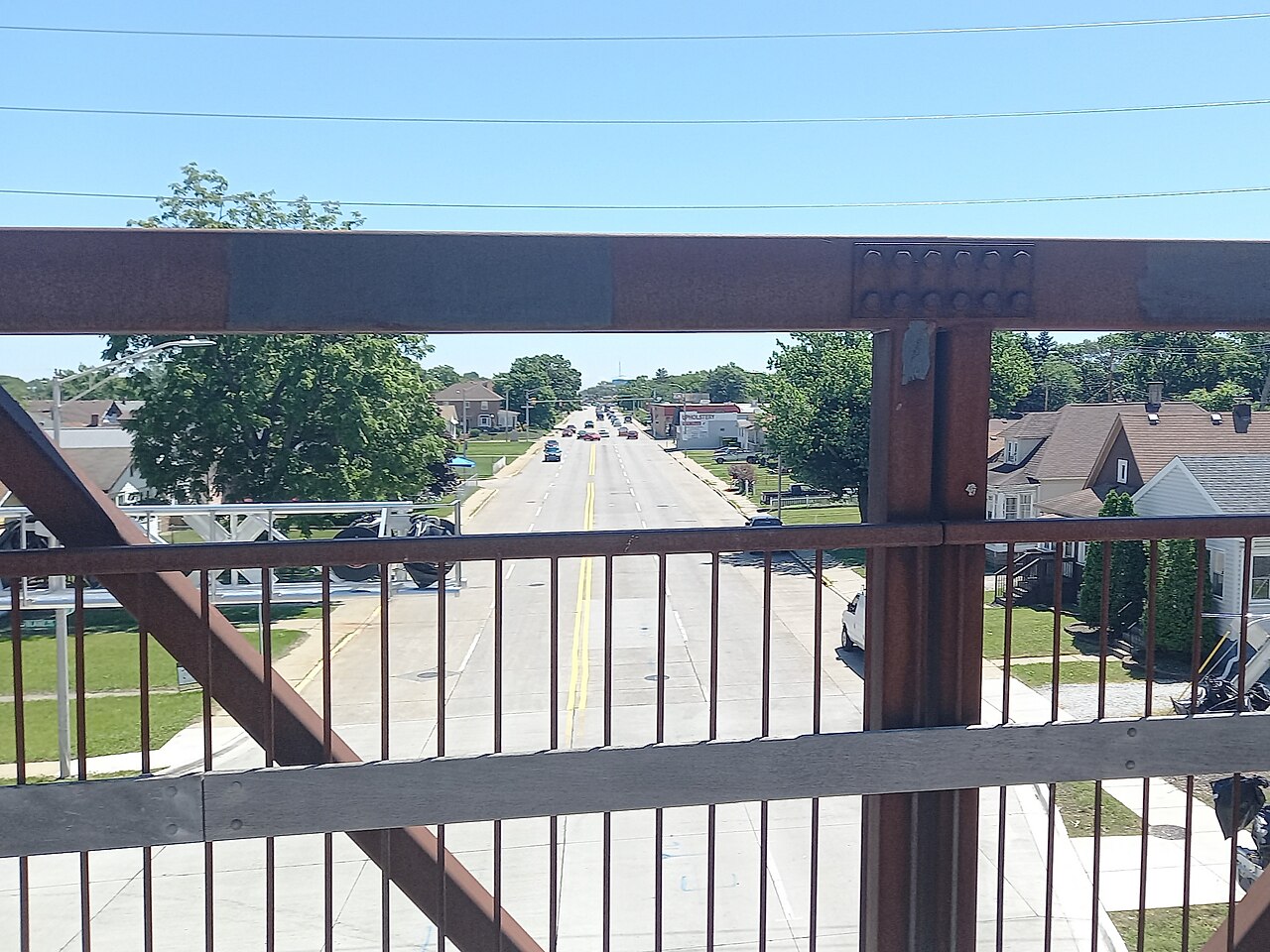 A wide two-way street with a double yellow center line stretches toward the horizon between houses and trees, seen through the rusted railing of a pedestrian bridge